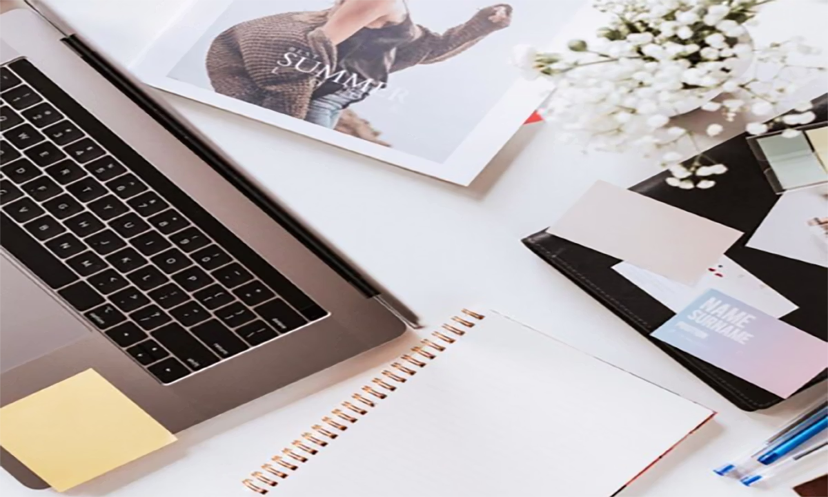 Overhead desk flat lay with a laptop, blank notebook, magazine page, white flowers, portfolio, and business cards.